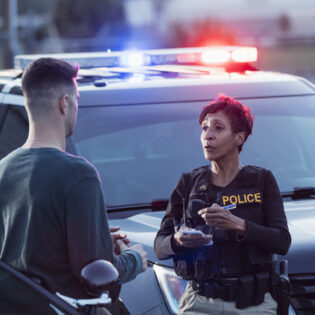 A policewoman taking a statement from a civilian outside her patrol car. The officer is a mature African-American woman in her 40s. She is talking with a young man in his 20s.